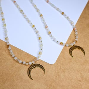 May include: Two delicate necklaces with gold crescent moon pendants. Each necklace features small white and light brown beads.