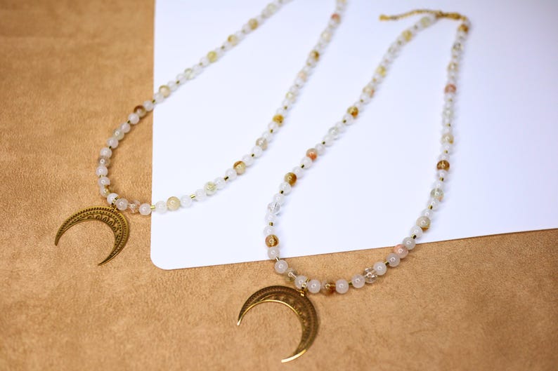 May include: Two gold crescent moon pendants on necklaces with white and light brown beads.