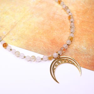 May include: A white and gold beaded necklace with a crescent moon pendant. The beads are small and round, and the pendant is made of metal with a detailed design.
