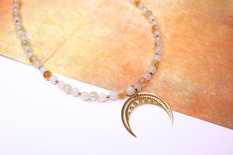 May include: A white and gold beaded necklace with a crescent moon pendant. The beads are small and round, and the pendant is made of metal with a detailed design.