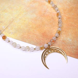 May include: A white and gold beaded necklace with a crescent moon pendant. The beads are small and round, and the pendant is detailed with intricate designs.