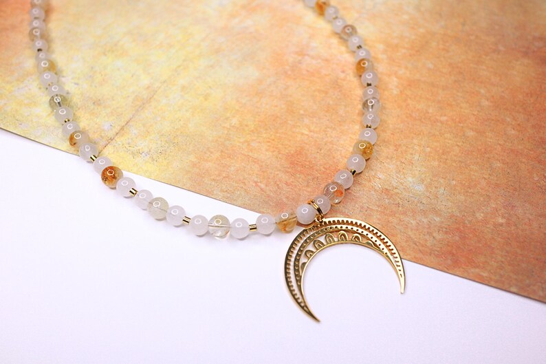 May include: A white and gold beaded necklace with a crescent moon pendant. The beads are small and round, and the pendant is detailed with intricate designs.