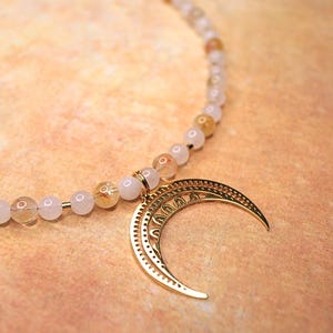 May include: A gold crescent moon charm hangs from a necklace made of white and gold beads.