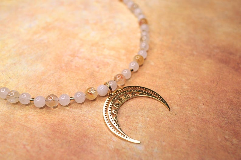 May include: A gold crescent moon charm hangs from a necklace made of white and gold beads.