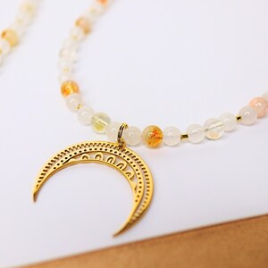 May include: A gold crescent moon pendant hangs from a necklace made of white and orange beads.