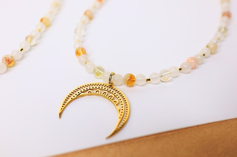 May include: A gold crescent moon pendant hangs from a necklace made of white and orange beads.