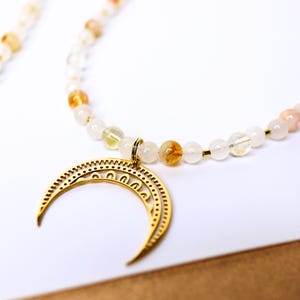 May include: A gold crescent moon charm hangs from a necklace made of white and orange beads.