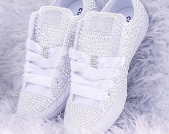 May include: White Converse sneakers with pearl embellishments and white satin ribbon laces. The sneakers have the Converse All Star logo on the side.