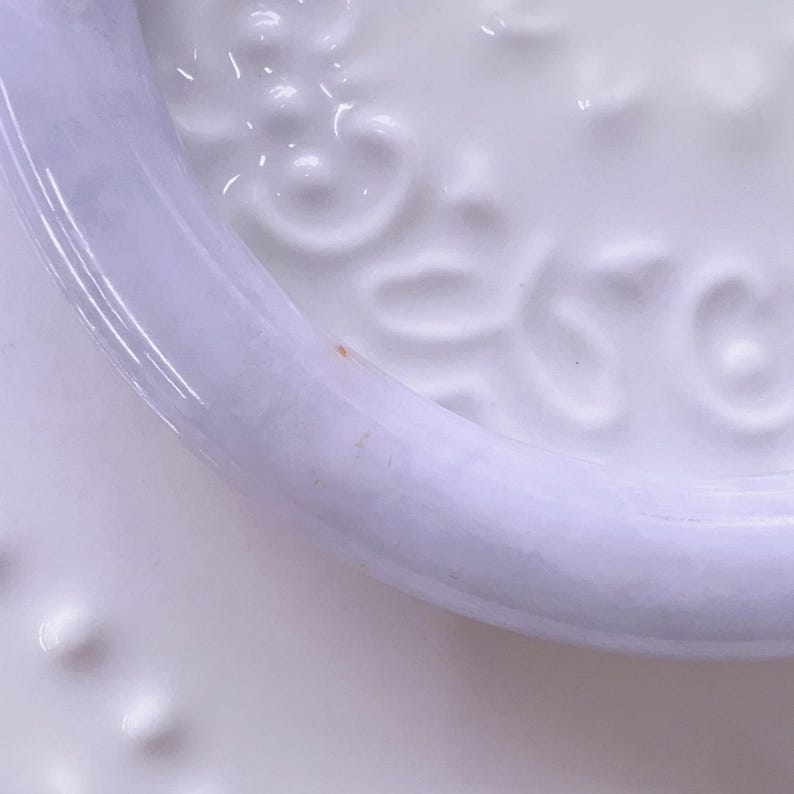 May include: A white ceramic plate featuring a raised, embossed floral pattern along the inner rim. The surface is smooth and glossy, with a distinct circular border separating the center from the edge. A small, faint orange speck is visible on the rim.
