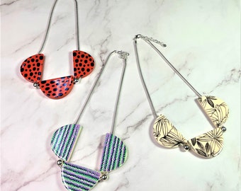 Op de afbeelding: Drie kettingen met zilveren kettingen en hangerontwerpen. Eén ketting heeft een rood-zwart polkadotpatroon, een andere heeft een groen-paars gestreept patroon en de derde heeft een crèmekleurige achtergrond met zwarte bloemmotieven.