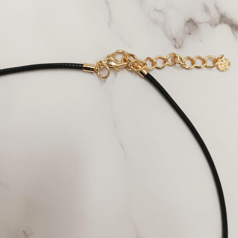 May include: Close-up of a black cord necklace with gold-coloured metal clasp and chain. The chain has a small decorative charm at the end. The necklace is set against a white marble background.
