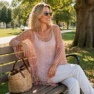 May include: A woman wearing a light pink, open-weave, fringed top over a white tank top and white pants. She is sitting on a wooden bench, wearing sunglasses, and holding a woven straw bag. The background shows a park with trees.
