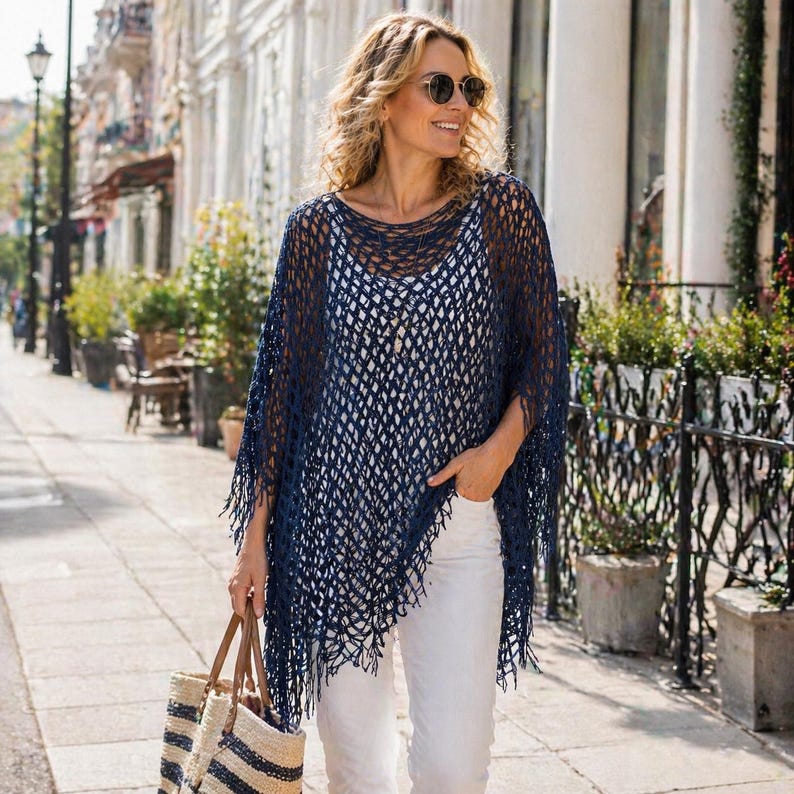 May include: A navy blue, open-weave poncho with fringe detailing. The poncho is worn over a white top and paired with white pants. A striped tote bag with brown leather handles is carried.