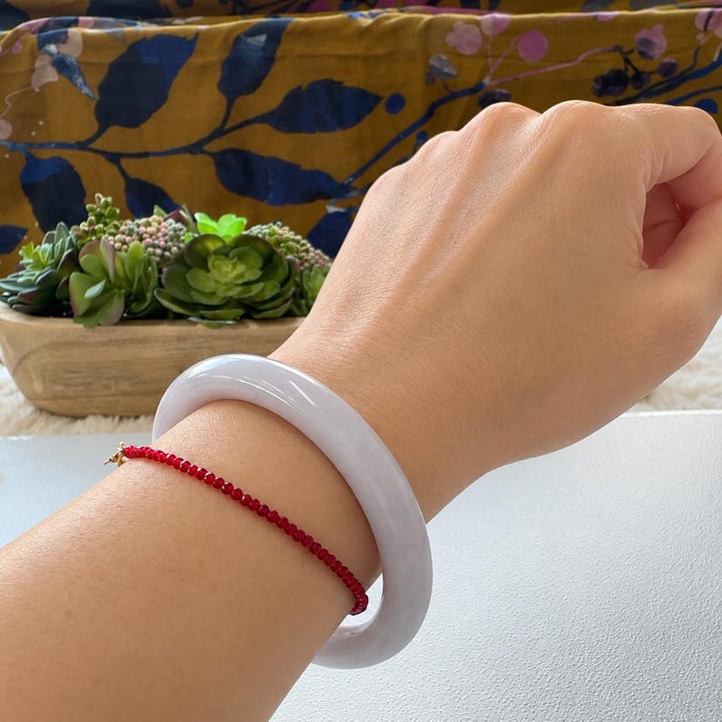 May include: A smooth, pale lavender-grey jade bangle bracelet with a rounded profile, worn alongside a delicate, thin bracelet composed of small, faceted, vibrant red beads on a fine gold-toned chain, creating a contrast in texture and color.