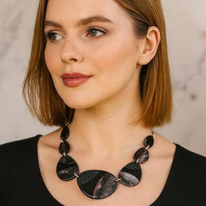 May include: A striking black necklace with a silver chain. The necklace has large, irregularly shaped pendants with a marbled black and purple design. The woman in the image is wearing a black top.