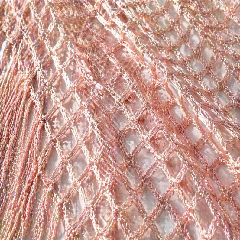 May include: Close-up of a shimmering, open-weave fabric in shades of rose and peach. The material has a diamond-shaped pattern, with a fringe detail along the edge. The texture appears soft and delicate, suitable for apparel or decorative use.