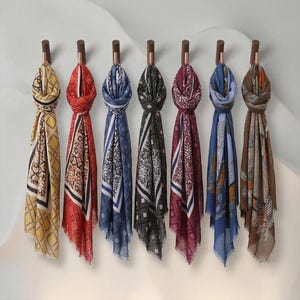May include: Seven rectangular scarves hanging on individual wooden wall hooks. Each scarf features a distinct color palette including yellow, red, blue, black, burgundy, light blue, and brown, all decorated with intricate geometric and floral patterns.
