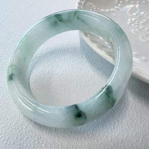May include: A smooth, polished bangle bracelet crafted from translucent pale green stone with darker green mottled inclusions throughout the circular band. The surface of the bangle is rounded and reflective, showcasing natural variations in the stone's color.