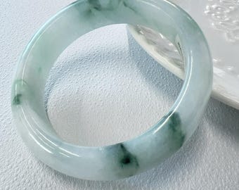 May include: A smooth, polished bangle bracelet crafted from translucent pale green stone with darker green mottled inclusions throughout the circular band. The surface of the bangle is rounded and reflective, showcasing natural variations in the stone's colour.
