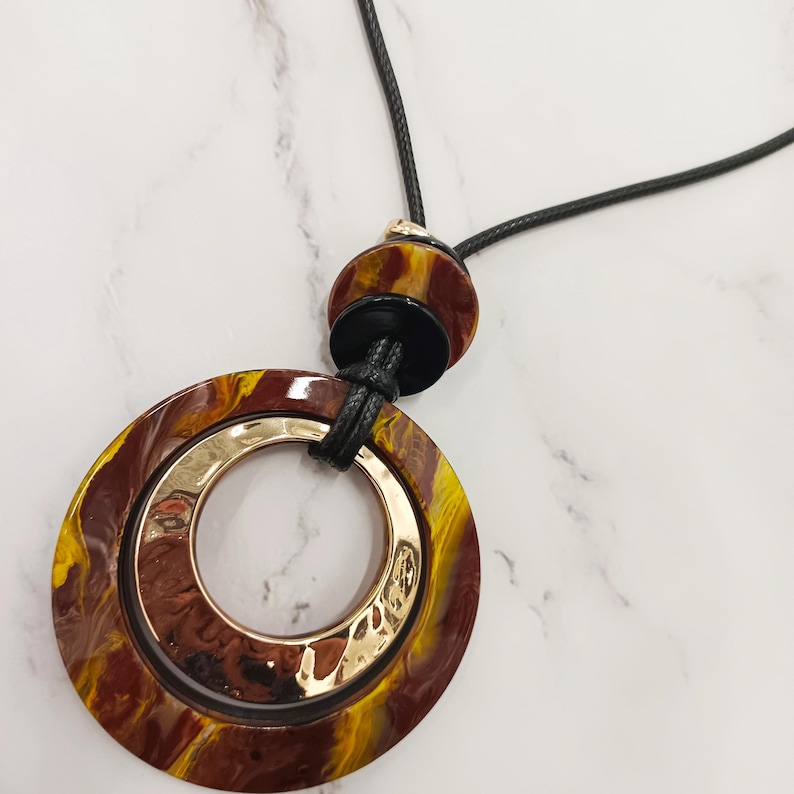May include: A statement necklace with a large, circular pendant. The pendant has a gold-coloured inner ring and a marbled outer ring in brown and yellow. It hangs from a black cord with a matching marbled bead.