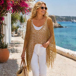 May include: A woman wearing a gold-colored, open-weave poncho with fringe. She is wearing a white top and white pants. She is carrying a straw tote bag with brown leather handles. The background includes a coastal scene with a blue ocean and white buildings.