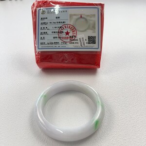 May include: A smooth, translucent white jade bangle bracelet featuring subtle green inclusions. The bracelet is accompanied by a red-wrapped authentication certificate that displays technical data, a QR code, and a photograph of the item.
