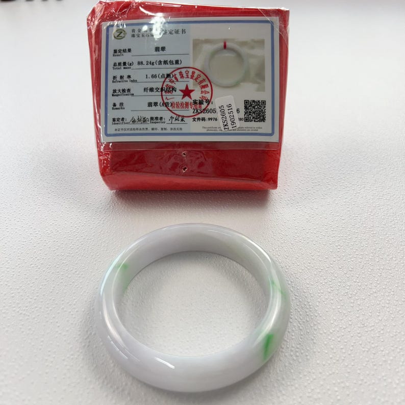 May include: A smooth, translucent white jade bangle bracelet featuring subtle green inclusions. The bracelet is accompanied by a red-wrapped authentication certificate that displays technical data, a QR code, and a photograph of the item.