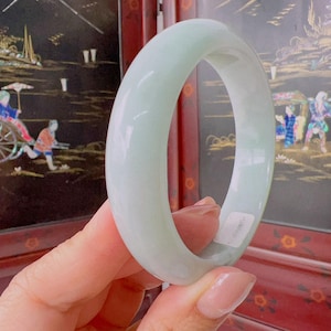 Op de afbeelding: Een gladde, gepolijste jade armband in een licht muntgroene kleur met een afgerond profiel. De massieve, cirkelvormige band vertoont subtiele variaties in kleur en doorschijnendheid over het hele oppervlak, met een klein wit plaklabel.