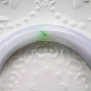 May include: A smooth, polished bangle bracelet crafted from translucent, pale lavender stone. The circular band features a distinct, vibrant green inclusion that creates a striking contrast against the soft, light purple base colour of the material.