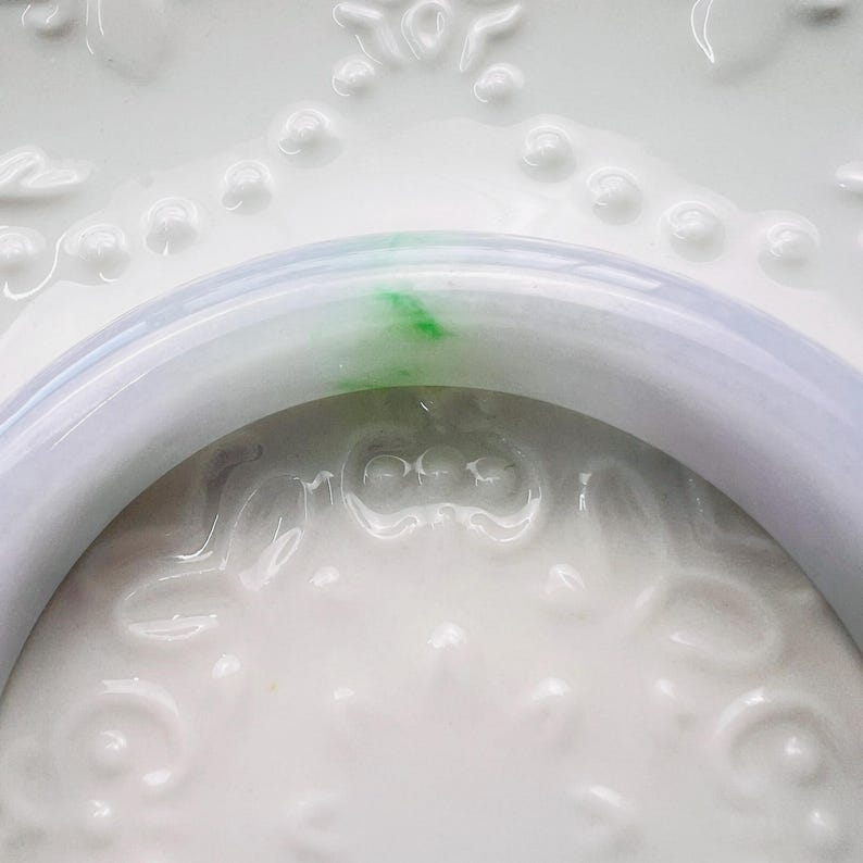 May include: A smooth, polished bangle bracelet crafted from translucent, pale lavender stone. The circular band features a distinct, vibrant green inclusion that creates a striking contrast against the soft, light purple base colour of the material.