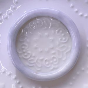 May include: A smooth, circular lavender-toned bangle bracelet with a polished finish. The bangle has a rounded profile and a consistent thickness throughout its circumference, showcasing a soft, light purple hue with subtle variations in color.