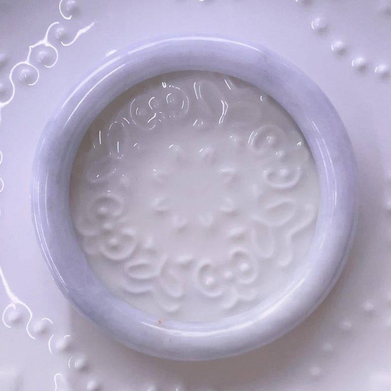 May include: A smooth, circular lavender-toned bangle bracelet with a polished finish. The bangle has a rounded profile and a consistent thickness throughout its circumference, showcasing a soft, light purple hue with subtle variations in color.