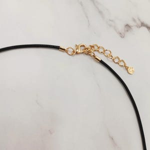 May include: Close-up of a black cord necklace with gold-coloured metal clasp and chain. The necklace has a small charm at the end of the chain. The background is a white marble surface.