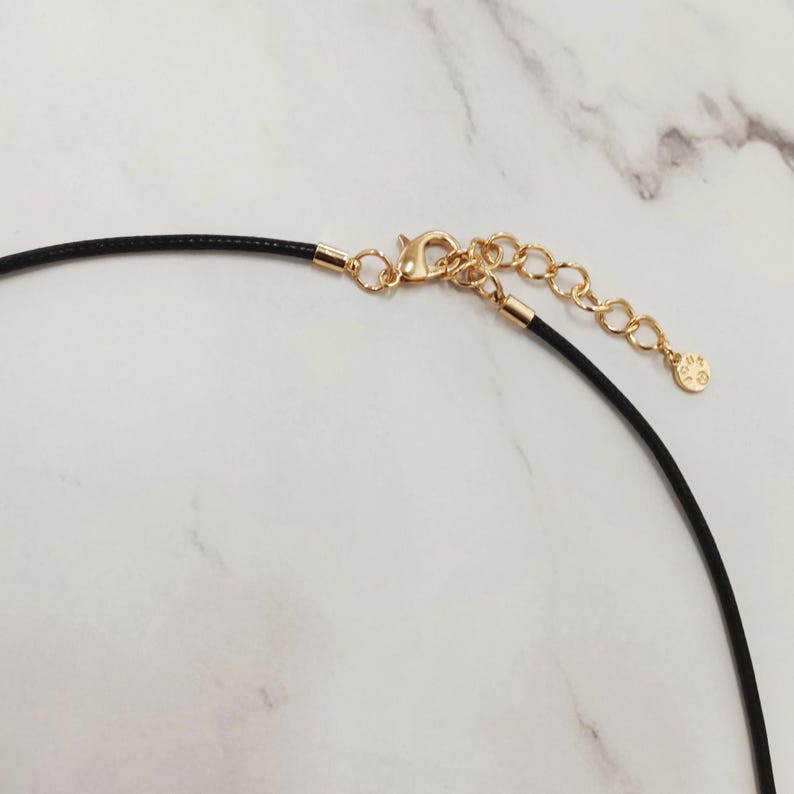 May include: Close-up of a black cord necklace with gold-coloured metal clasp and chain. The necklace has a small charm at the end of the chain. The background is a white marble surface.
