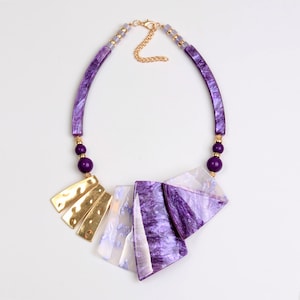 May include: A purple and gold statement necklace with a geometric design. The necklace features a large, multi-faceted pendant made of purple and white resin, with gold accents. The pendant is attached to a chain of purple beads and gold links.