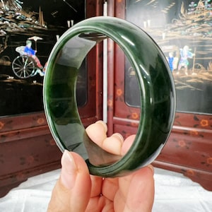 May include: A smooth, polished, dark forest green jade bangle bracelet with a rounded exterior and a flat interior surface. The material is translucent, showing variations in deep green colour and natural internal patterns throughout the band.
