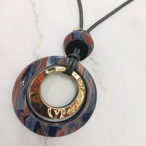 May include: A pendant necklace with a large, marbled blue, red, and gold circular pendant. The pendant has a gold inner ring and is suspended from a black cord. The necklace has a matching marbled bead at the top.