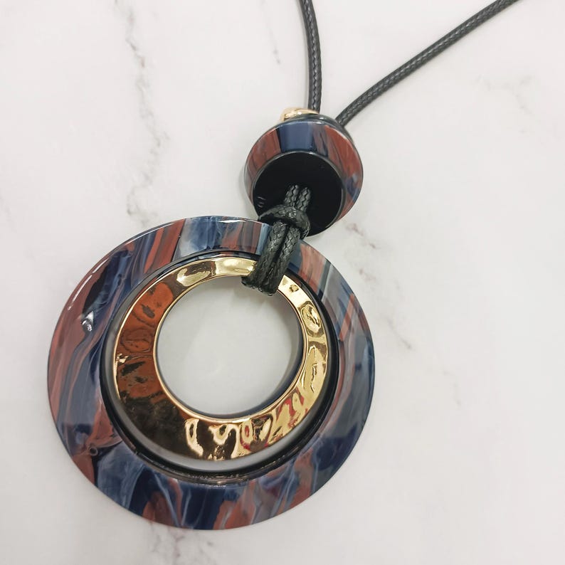 May include: A pendant necklace with a large, marbled blue, red, and gold circular pendant. The pendant has a gold inner ring and is suspended from a black cord. The necklace has a matching marbled bead at the top.