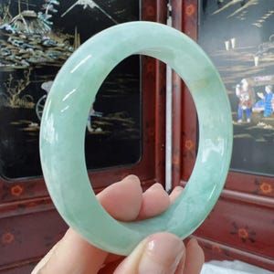 May include: A smooth, polished, light mint green jade bangle bracelet with a rounded profile. The stone features subtle, translucent variations in colour and texture throughout its circular form, creating a soft, glossy appearance.