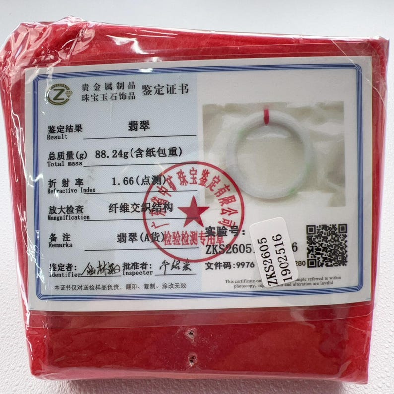 May include: A white jade bangle inside a clear plastic bag with a red fabric backing. A certificate of authenticity is attached, displaying a total mass of 88.24g, a refractive index of 1.66, and a QR code with the serial number ZKS26051902516.
