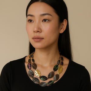 May include: A two-strand necklace with oval beads in brown, grey, and iridescent green and purple. Gold-coloured, patterned oval accents are interspersed. The necklace is worn by a person wearing a black top.