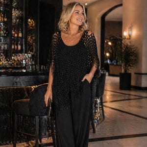 May include: A woman wearing a black crochet top with fringe and a black jumpsuit. The top has a scoop neck and open weave design. The jumpsuit has wide legs and pockets. The background includes a bar and chairs.