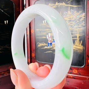 May include: A smooth, circular bangle bracelet crafted from translucent white jadeite with subtle, vibrant green inclusions. The bangle features a polished, rounded surface and a solid, continuous band design with a glossy, reflective finish.