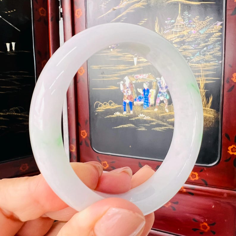 May include: A smooth, translucent white jade bangle bracelet featuring subtle, pale green inclusions throughout the polished stone surface. The circular jewellery piece has a rounded profile and a glossy finish, showcasing natural colour variations.