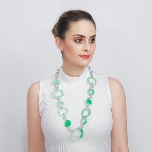 May include: A green and silver necklace with a variety of geometric shapes. The necklace has a silver chain and a series of green, translucent, and opaque circular and polygonal pendants. The necklace is worn by a person wearing a white top.