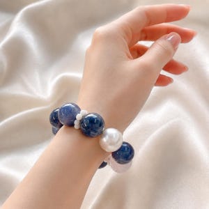 May include: A beaded bracelet featuring large, round, dark blue mottled beads interspersed with a single large, smooth, glossy white bead and smaller, opaque white spacer beads. The bracelet is constructed with a flexible, elasticised cord design.