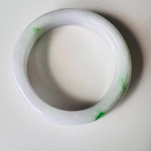 May include: A smooth, circular bangle bracelet crafted from polished stone. The material is primarily a translucent, pale lavender-white colour, featuring distinct, vibrant green mineral inclusions that appear as small, irregular patches.