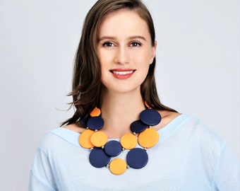 May include: A woman wearing a light blue shirt with a necklace made of large orange and dark blue circles.