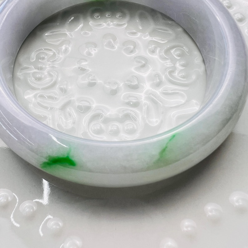 May include: A smooth, polished jade bangle bracelet featuring a translucent light lavender hue with distinct, vibrant green inclusions. The circular band has a rounded profile and a glossy finish that reflects light across its curved surface.