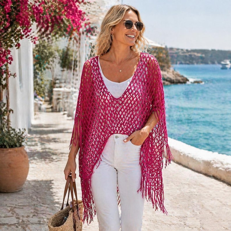 May include: A magenta-colored, open-weave poncho with fringe detailing. The poncho is worn over a white top and white pants. The image is taken outdoors with a coastal background.